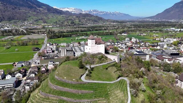 balzers village rooftops modern buildings hillside gutenberg castle hilltop stone tower drone aerial orbit alpine peaks mountain range green valley farmland liechtenstein rhine valley heritage site