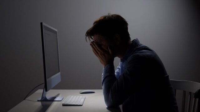 Exhausted man covering his face at a dim computer desk in a dark office, conveying work burnout, late night stress, overwhelm, fatigue and emotional pressure under deadline