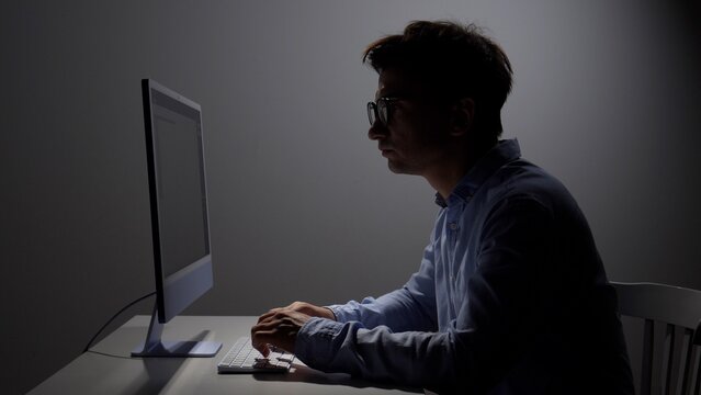 Man in profile wearing glasses concentrates on laptop at a dim desk, screen glow highlighting his face and hands as he types late into the night, focused on coding or work tasks