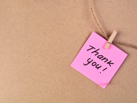 Thank you handwritten text on a square pink paper note, clipped with a wooden clothespin to a rustic jute string on a brown cardboard background. Ideal for customer appreciation concept