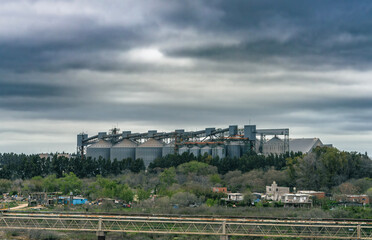 Industrial grain terminal facility with large metallic storage silos and conveyor systems set behind green trees and a foreground bridge under a dramatic overcast sky near a waterway © John_Doo78