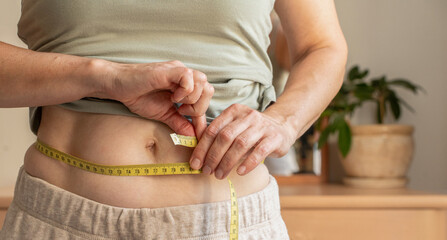 Close-up, woman measuring her waist with yellow tape measure at home. Concepts include weight loss...