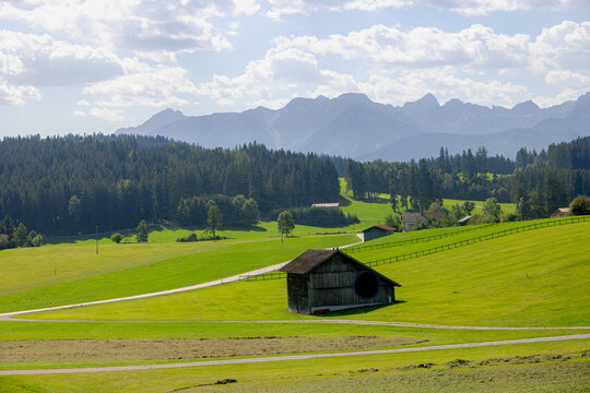 Rural Bavarian countryside landscape, Traditional wooden houses and barn on rolling hills, Lush green grass meadow against majestic Alpine mountains under blue sky and white clouds in Bavaria, Germany