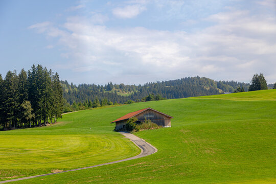 Bavarian countryside landscape, A traditional wooden barns standing alone on a rolling hill, Lush green grass meadow against majestic Alpine mountains under blue sky and white clouds, Bavaria, Germany