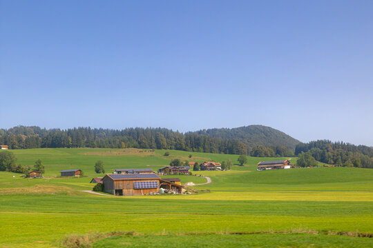 Rural Bavarian countryside landscape, Traditional wooden houses and barn on rolling hills, Lush green grass meadow against majestic Alpine mountains under blue sky and white clouds in Bavaria, Germany