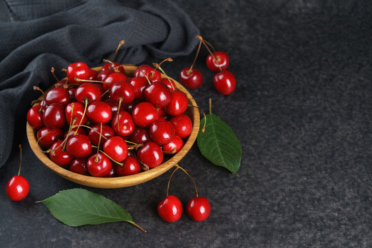 Fresh ripe red sweet cherries in wooden bowl on dark textured stone background with green leaves and black linen towel. Summer fruit, berry, harvest, healthy organic food