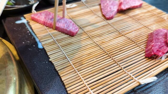 Chopsticks placing slices of raw beef on a bamboo mat, showcasing wagyu meat with a marbled fat pattern, slow motion footage.