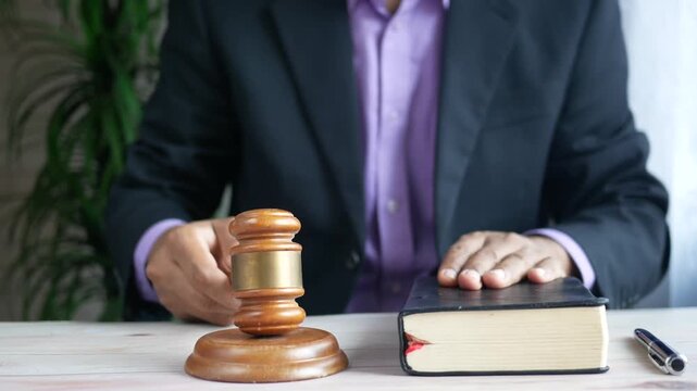 Unrecognizable judge striking gavel on sound block and holding bible in courtroom setting during legal proceeding