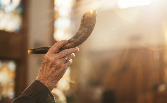 Elderly man holding a traditional shofar horn in sunlight, representing faith, spirituality, and Jewish holiday rituals indoors.