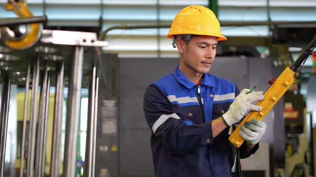 Asian male industrial worker wearing hard hat and uniform holding remote controller in manufacturing plant, Factory engineer operating heavy machinery with crane remote control in industrial facility