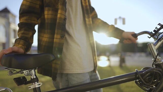 Bike city bicycle transport. A man in a plaid shirt holds a bicycle seat while near a bicycle. The man stands on grass with sunlight shining in the background near a park.