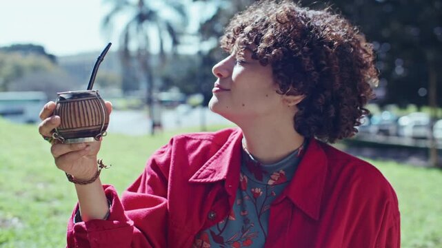 Portrait of cheerful young woman with curly hair drinking mate tea and smiling at camera, then posing with traditional calabash gourd outdoors in park