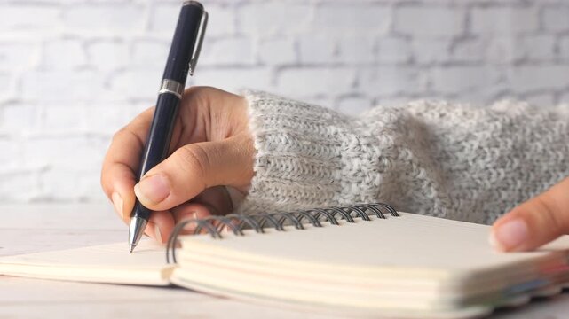 Person writing in spiral notebook with black pen on white brick wall background
