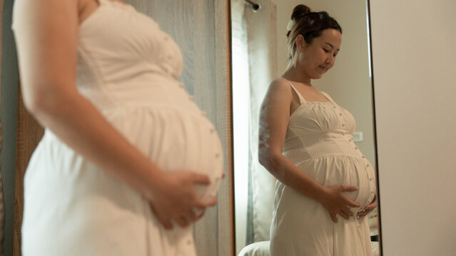 Close-up of a pregnant woman in a white dress looking down tenderly at her belly, reflected in the mirror.