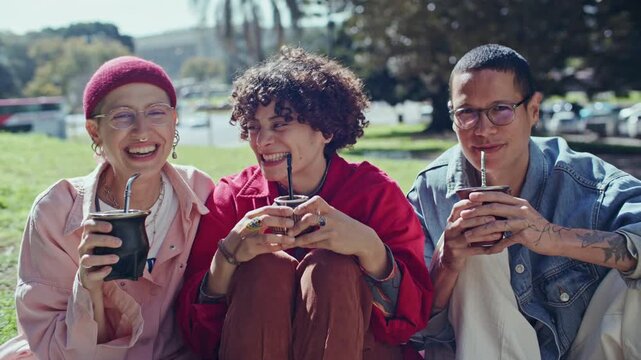 Diverse group of three joyful young friends laughing and smiling at camera while enjoying traditional South American yerba mate tea in city park. Video portrait