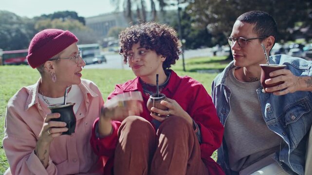 Group of three young multiethnic friends sitting on grassy hill in public park, talking and laughing while drinking traditional yerba mate tea from gourds on sunny summer day