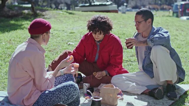 Curly-haired girl pouring hot mate tea from thermos into calabash gourd cups for her friends as enjoying outdoor picnic in park on sunny day