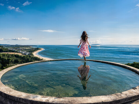 The a little girl stands in a pool, facing the vast expanse of the sunny ocean and tropical beach.