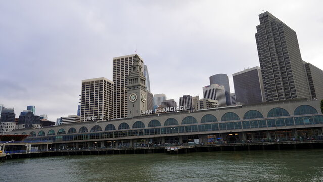 The iconic arched facade and landmark clock tower of the San Francisco Ferry Building sit along the Embarcadero waterfront, backed by the skyscrapers of the downtown Financial District under an overca