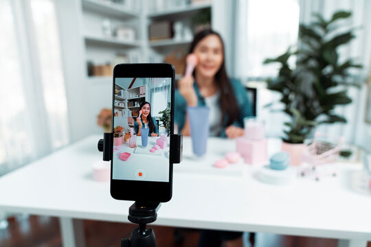 Selective focus smartphone with young beautiful Asian reviewing cosmetic makeup brush cheek test collection with pastel color, showing glow blusher mood to promote on social media online. Stratagem.