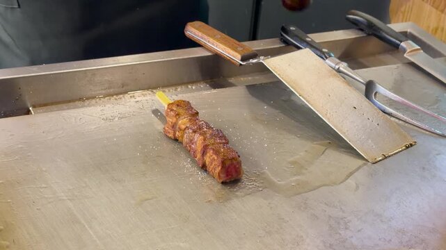 Chef seasoning a delicious meat skewer on a hot griddle with salt and pepper in a dynamic close up shot.