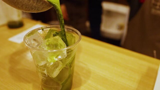 Pouring fresh organic matcha green tea over ice cubes in plastic cup.