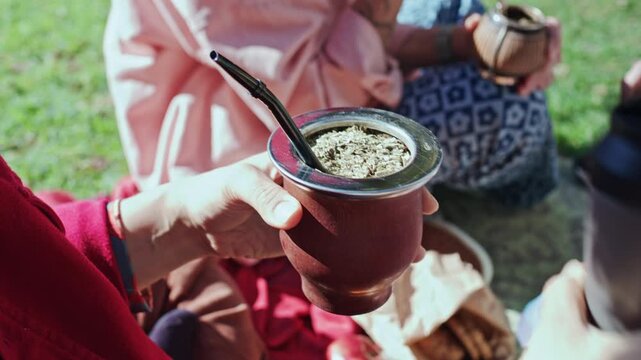 Close up shot of female hands pouring hot water from thermos into traditional ceramic mate gourd filled with organic loose yerba mate leaves during park picnic