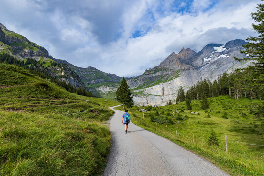 A young man in a backpack hikes along a paved path toward the mountains over Oeschinen Lake (Oeschinensee) near Kandersteg, Switzerland