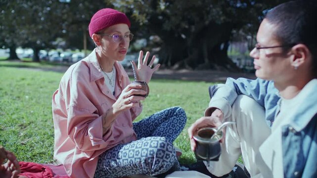 Young woman sitting on grass, chatting with friend and drinking traditional South American yerba mate tea in city park on sunny day