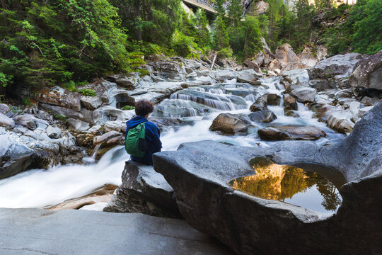 A young man sits along the Avers Rhine river in the Magic Wood climbing area of Ausserferrera, Switzerland. In the foreground a small pool of water reflects golden light reflected from rock face above