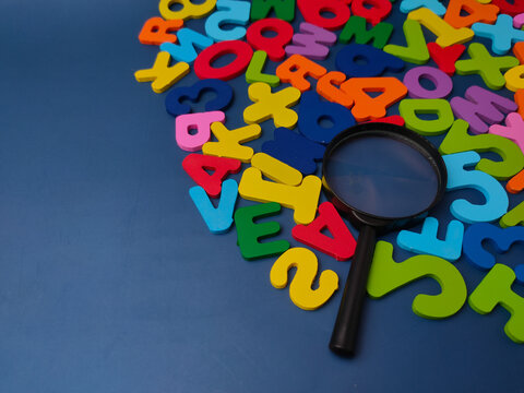 A black magnifying glass lies on a collection of colorful wooden letters and numbers over a blue surface
