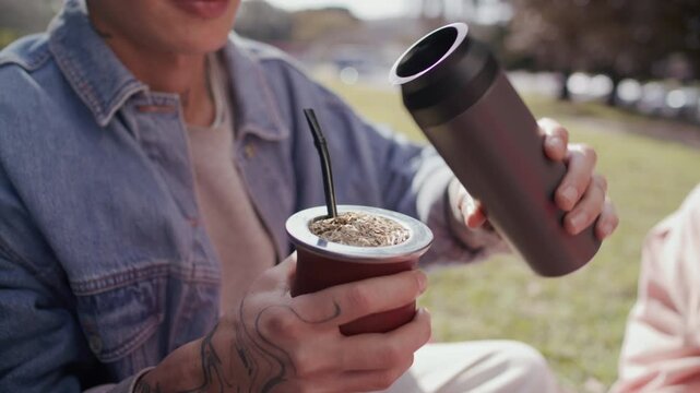 Young man pouring hot water from thermos into traditional yerba mate gourd and drinking tea through straw during outdoor picnic in park. Close-up shot