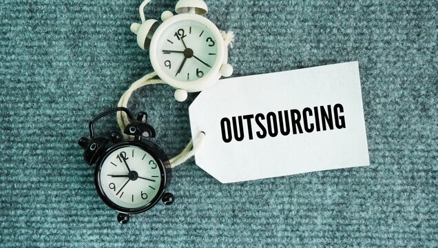Bell clocks and paper tags with the words Outsourcing is the business practice of hiring an external party or outside company to handle tasks, services, or production that a company would normally 