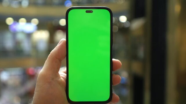 Hand holds smartphone with green screen. Blurred mall background features lights and moving figures. Green screen acts as placeholder for digital content. Ideal for screen recording demonstrations
