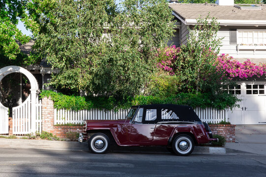 Los Angeles, California, USA - September 12, 2026: Vintage burgundy car parked along residential street beside landscaped home, white fence, brick wall, trees, and vibrant pink flowers.