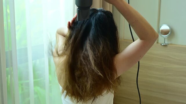 Rear view of Asian woman using hair dryer for blows hot air to speed the evaporation of water to dry the hair in her bedroom.