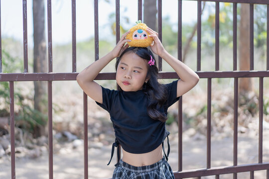 Portrait cute little girl holding plush toy on head black shirt plaid skirt metal gate outdoor playful youth childhood innocent fun lifestyle expression happy kid