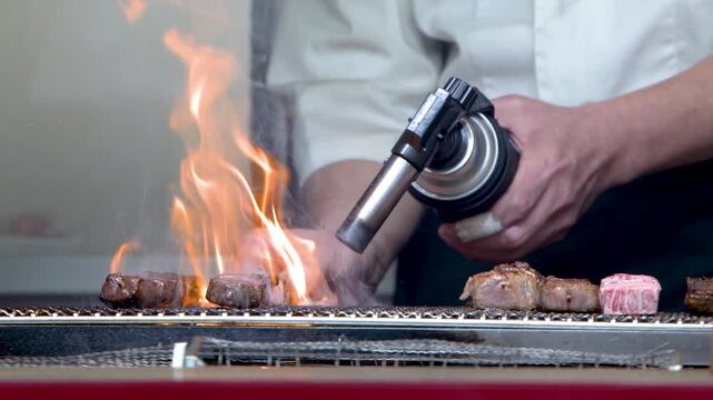 Chef searing cubes of meat on grill with culinary torch for delicious food preparation.