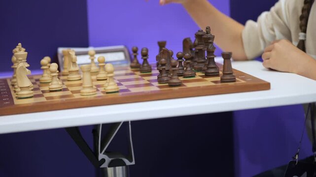 Chess game in progress on wooden board. Player moves piece with focused hand. Digital timer visible beside board. Blue background adds contrast. White table supports setup