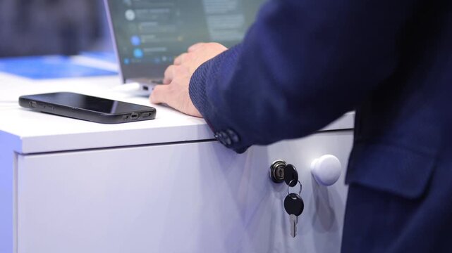 Person in dark suit uses laptop on white desk. Smartphone lies beside laptop. Keys hang from drawer handle. Background shows blurred tech display