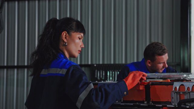 Industrial worker protective clothing and blue safety uniform inspects EV battery equipment inside metal garage, focused attention, careful teamwork, technical skill, and maintenance dim industrial