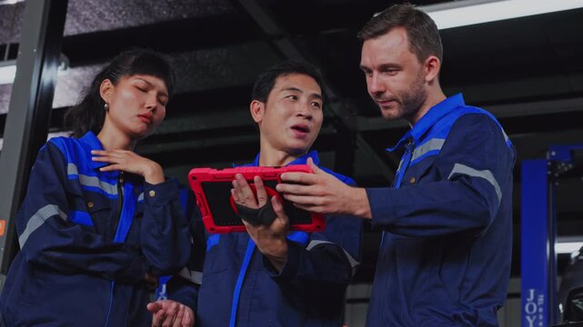 Technical workers blue uniform reviewing tablet beside EV vehicle inside garage workshop, industrial teamwork during automotive service training, focused collaboration and professional inspection