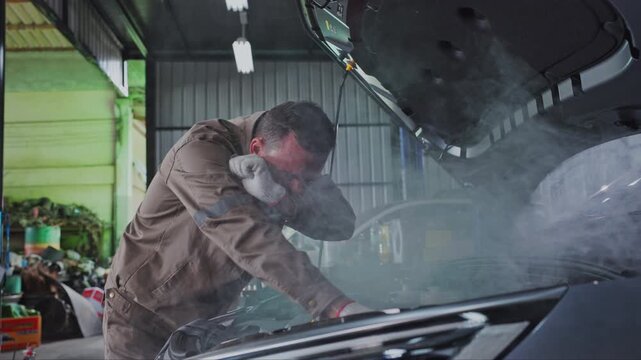 Person repairing car in smoky garage with open hood, mechanic inspecting smoking engine and visible EV components during focused vehicle repair and maintenance work with visible concern