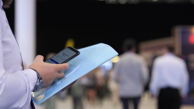 Person holds smartphone and blue folder at night event. Background shows blurred attendees walking. Scene is indoors with artificial lighting. Focus remains on hands and objects