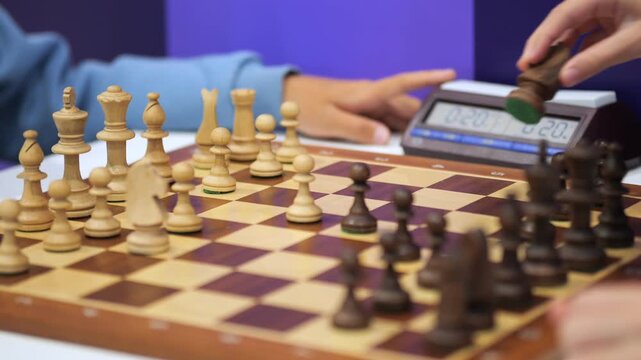 Close-up of chess players' hands moving pieces on wooden board. Two players focused during game. Digital clock visible beside board. Game in progress with pieces arranged for next move