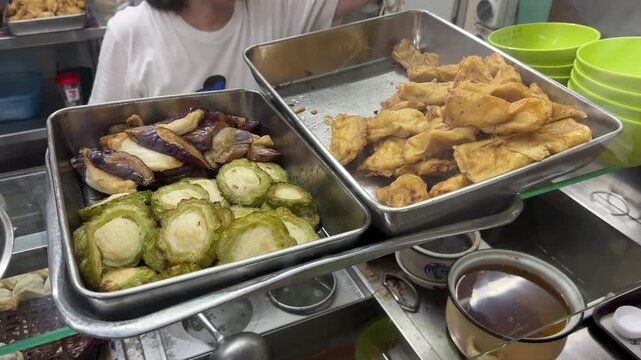 Yong tau foo ingredients displayed at a hawker stall, featuring stuffed eggplant, bitter gourd, and golden fried tofu puffs on metal trays, ready to be selected and cooked. Singapore street food