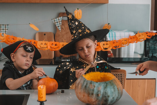 A portrait of a young girl and her younger brother celebrating Halloween and carving pumpkins in the kitchen with their mother's help. They are dressed in witch and pirate costumes. 