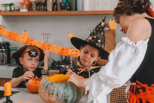 A portrait of a young girl and her younger brother celebrating Halloween and carving pumpkins in the kitchen with their mother's help. They are dressed in witch and pirate costumes. 