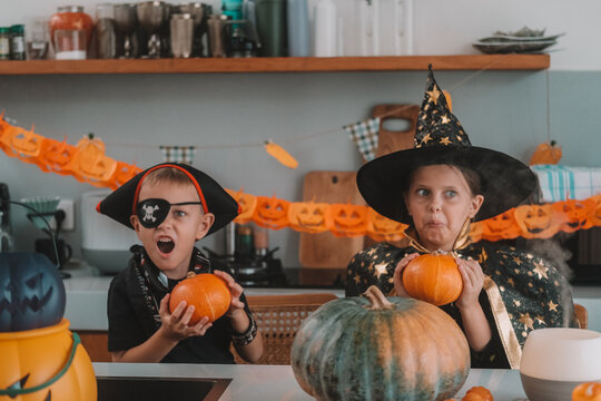Portrait of a girl and her a little brother celebrating Halloween and decorating pumpkins in the kitchen. They look scary in witch and pirate costumes. Kids Halloween party.