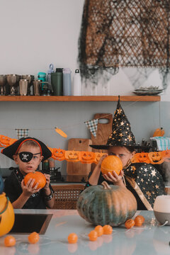 Portrait of a girl and her a little brother celebrating Halloween and decorating pumpkins in the kitchen. They look scary in witch and pirate costumes. Kids Halloween party.